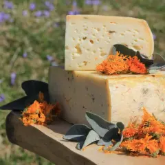 Due blocchi di formaggi di montagna, uno sopra l'altro, decorati con fiori arancioni e foglie verdi su un tagliere di legno.
